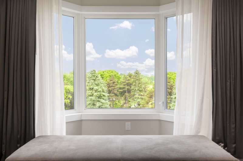 An interior view of a set of bay windows. A black and white curtain surrounds the outside, and a blue sky and trees can be seen outside.