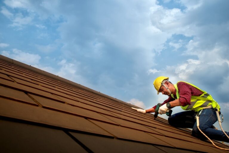 A roofing contractor in protective gear installs brown shingles onto a roof. You can see a blue, cloudy sky in the background.